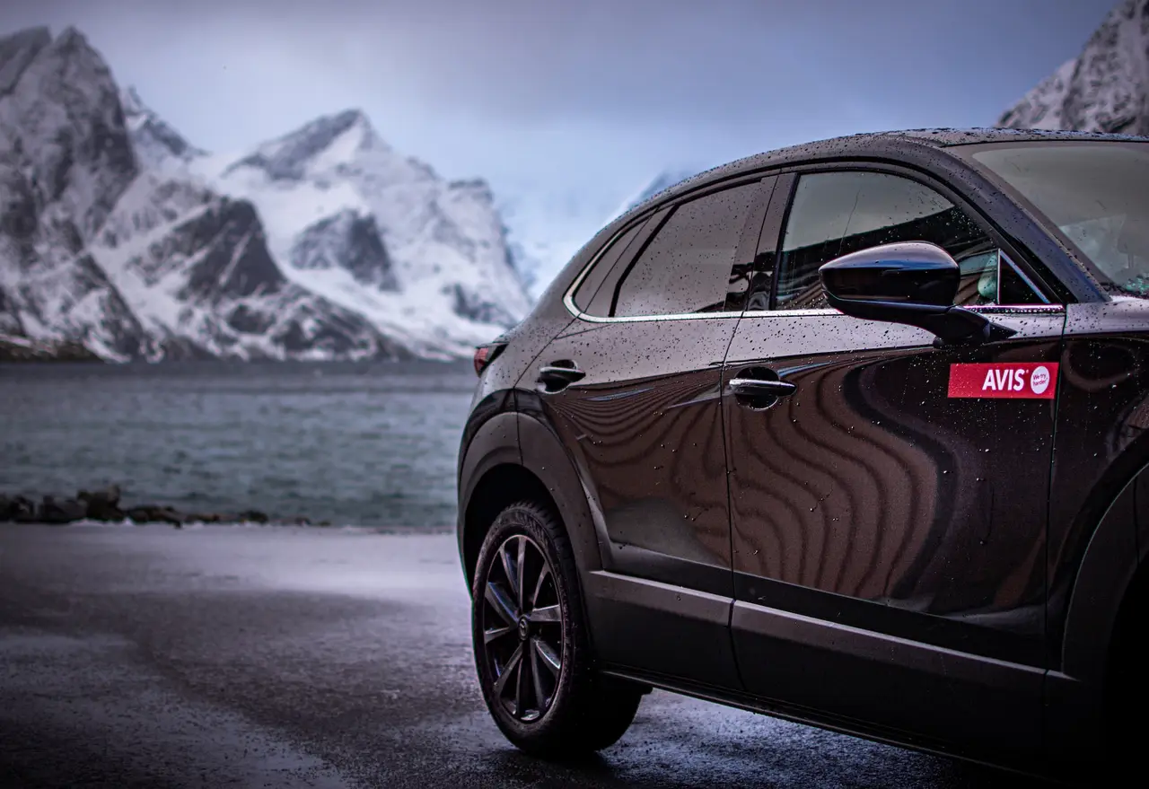 An Avis hire car parked on a road near water and snowy Norweigan mountains.