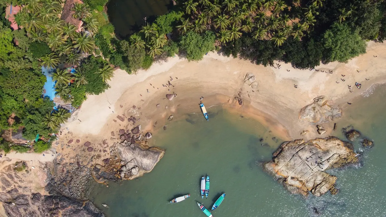 An aerial photograph of Gokarna's Paradise Beach, taken during the daytime. Beachgoers and boats can be seen amongst rocks, trees, and sand.